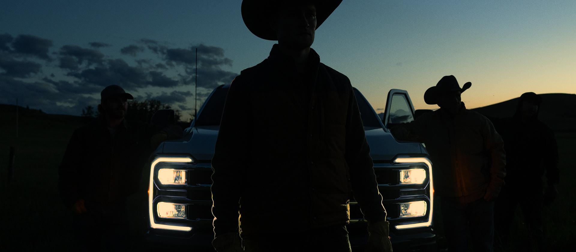 Silhouette of a person in a cowboy hat standing in front of a 2027 Ford Super Duty® truck with the headlights on at dusk