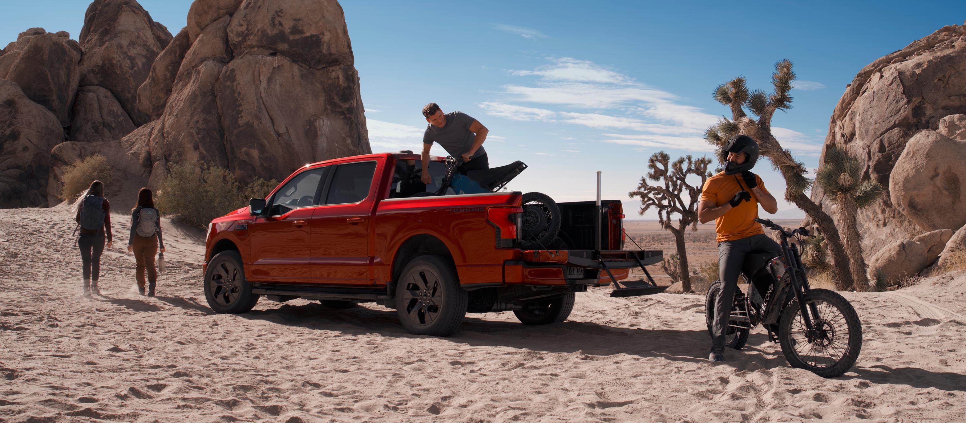 Le F-150 Lightning 2025 stationné sur du sable avec des roches et des cactus en arrière-plan. Un groupe de personnes partant en randonnée