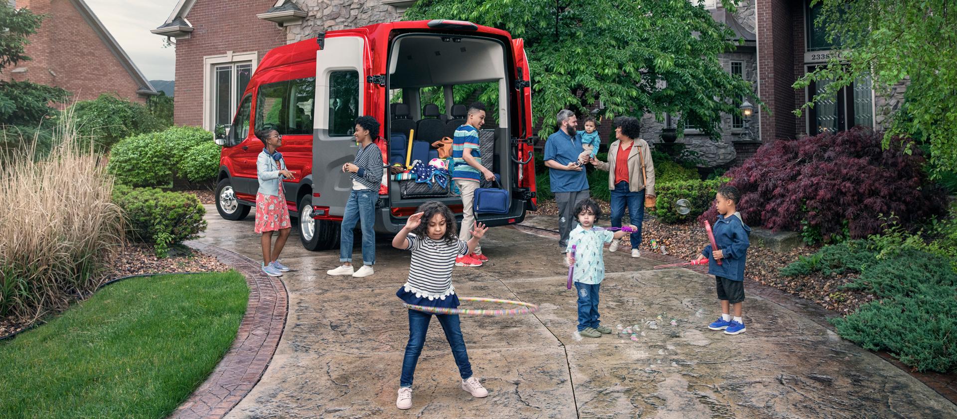 Une grande famille réunie dans une entrée de garage de banlieue autour d’une fourgonnette de transport de passagers Ford Transit® rouge aux portes ouvertes, avec des enfants qui jouent au premier plan