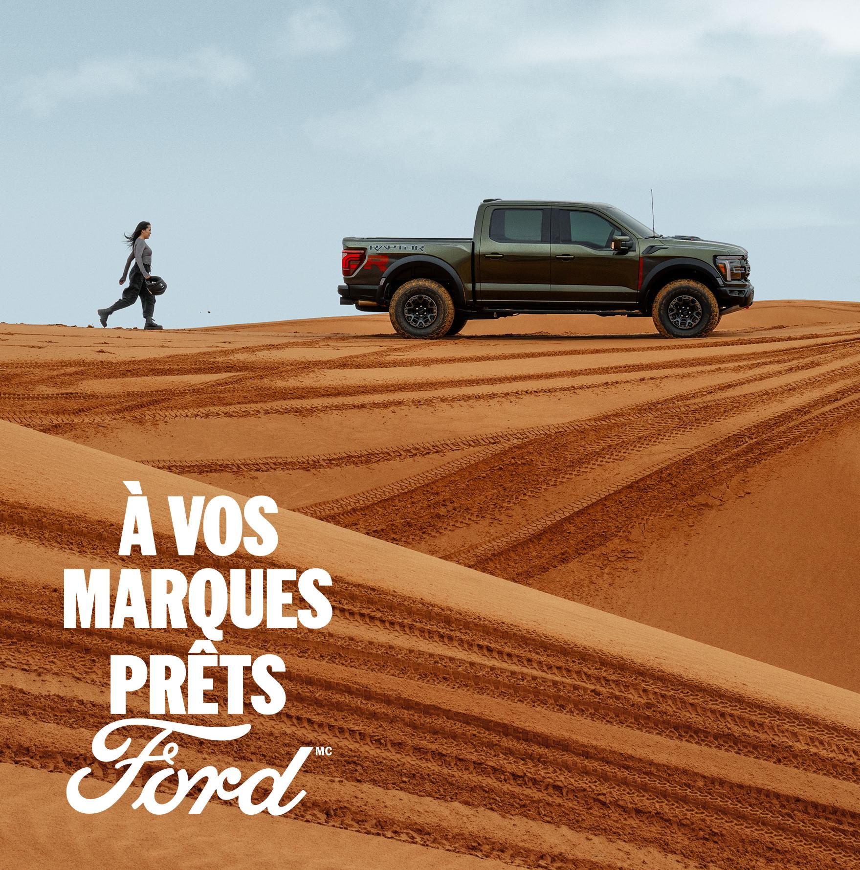Une femme marche vers l'arrière d'un Ford F-150 Raptor gris, vu de profil, dans un paysage désertique.