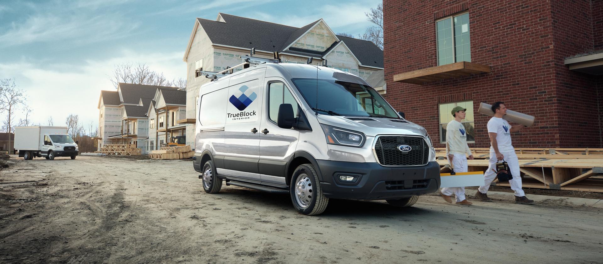 Fourgonnette Ford Transit® 2027 blanche équipée d’un support d'échelles sur un chantier de construction, avec deux ouvriers transportant des matériaux