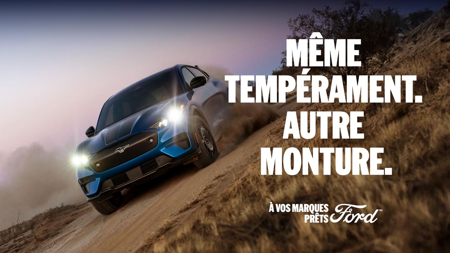 Vue de trois quarts avant d'un SUV électrique Ford Mustang Mach-E bleu roulant en biais sur un chemin de terre. 