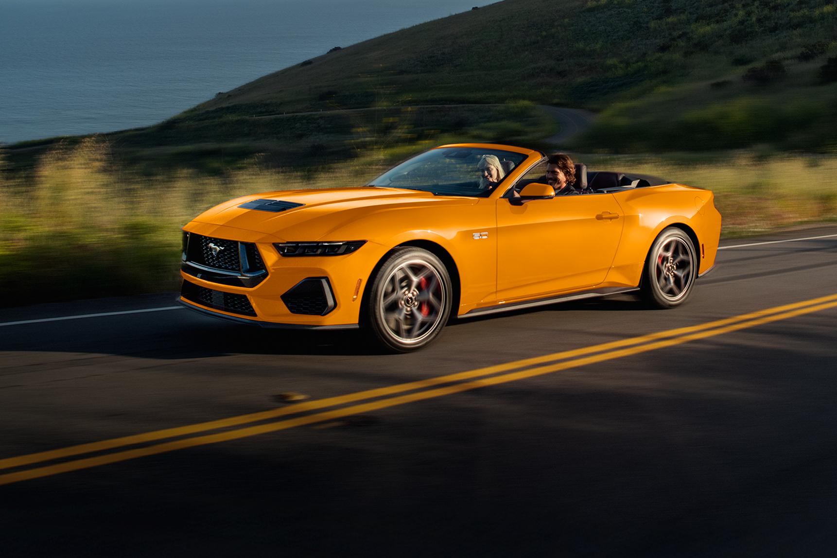 Deux personnes à bord d'une Ford Mustang® GT décapotable 2026 de couleur Orange furie métallisé à trois couches roulant sur une route côtière