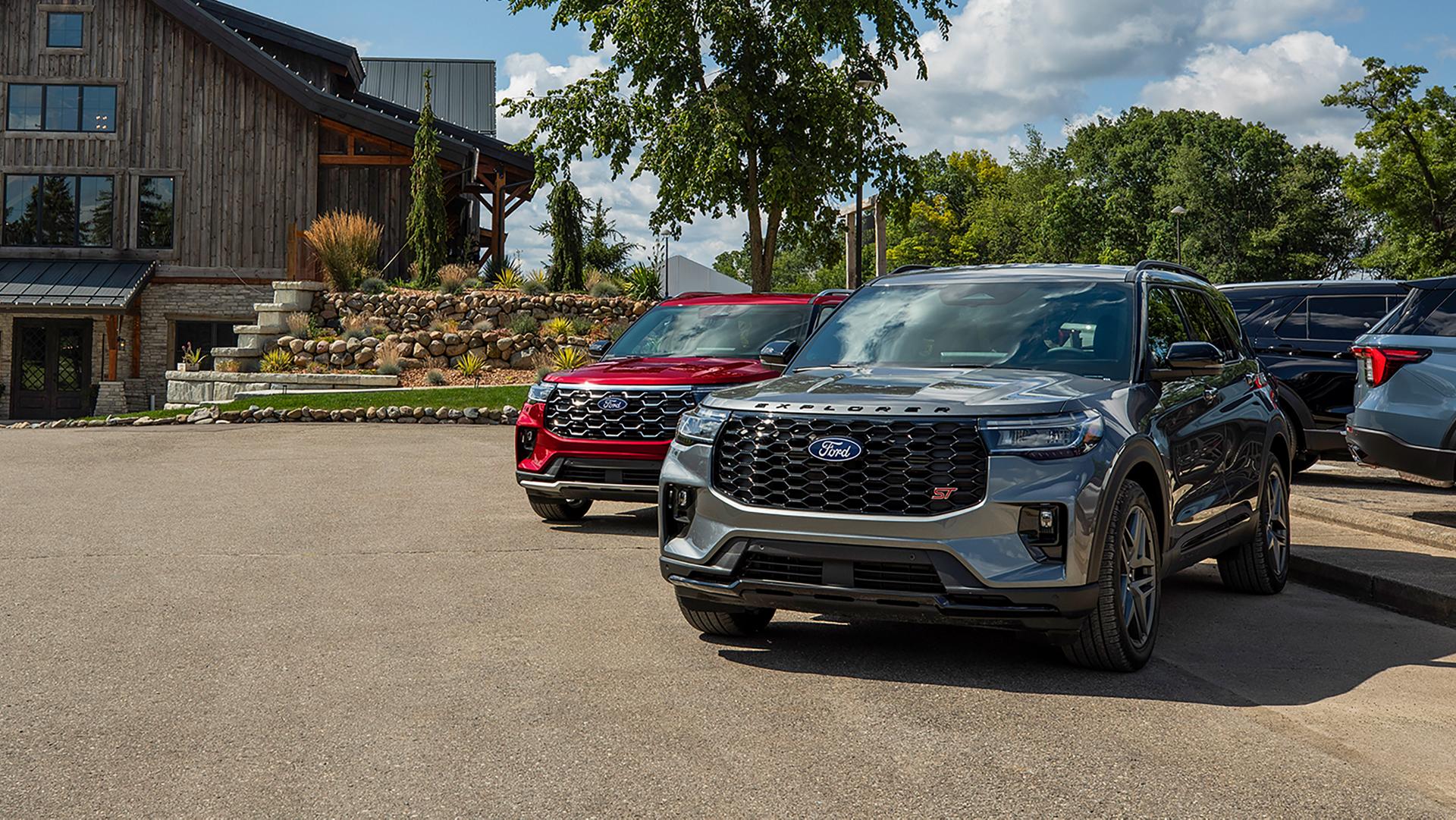 Deux VUS Ford Explorer® 2027 stationnés côte à côte dans un stationnement près d’arbres et d’un bâtiment rustique