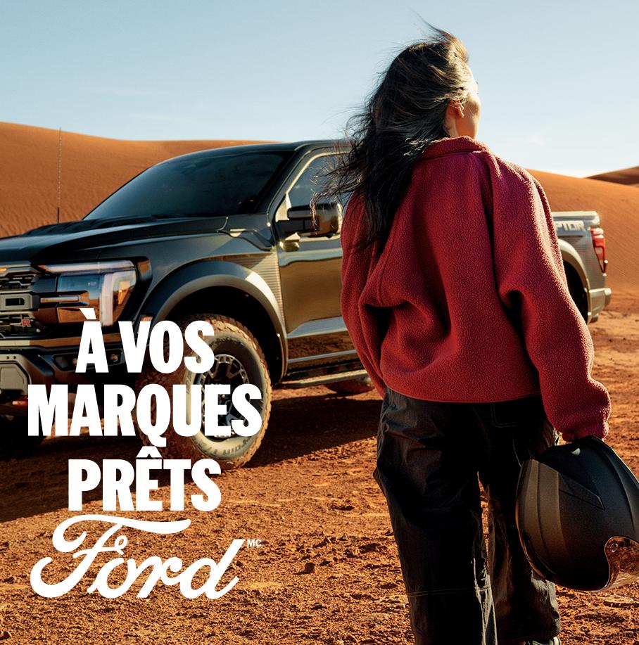 Une femme, dos tourné, tient un casque alors qu'elle est debout dans le désert devant un Ford F-150 Raptor gris foncé. Le mot « Ford » en blanc dans le coin gauche. Le texte blanc « À vos marques. Prêts.Ford » dans le coin inférieur droit