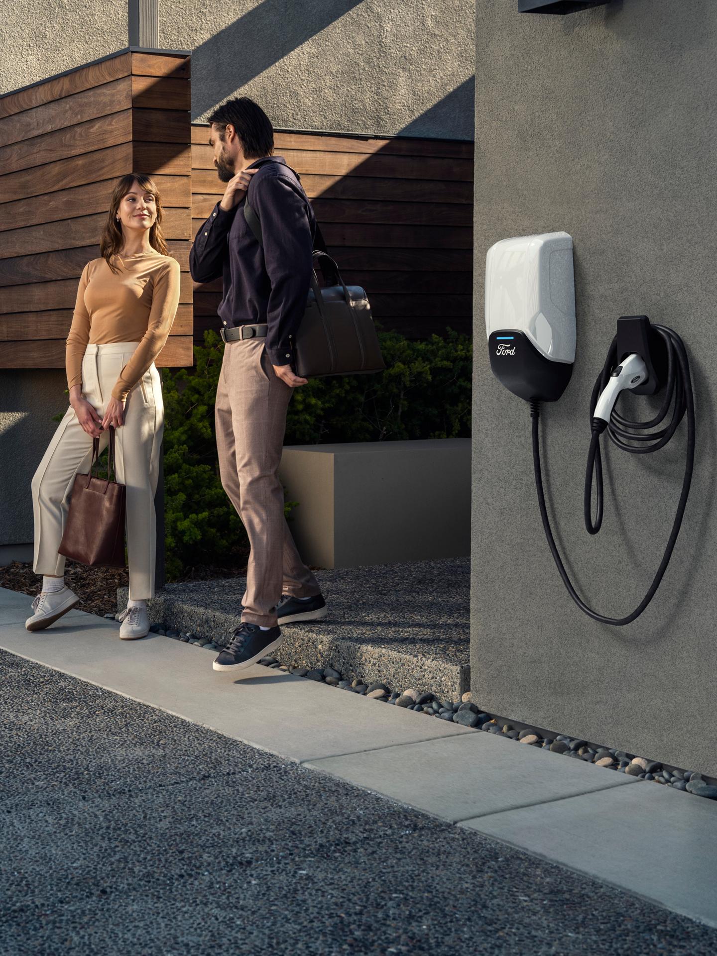 Un couple discute devant une maison où une borne de recharge Ford Pro est installée sur un mur à proximité.