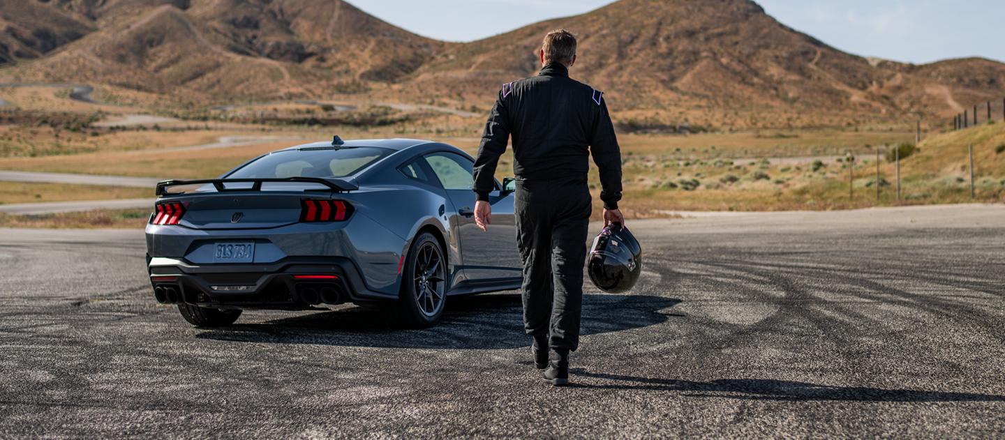 Une personne tenant un casque de course automobile marche vers une Mustang Dark Horse stationnée sur un circuit.