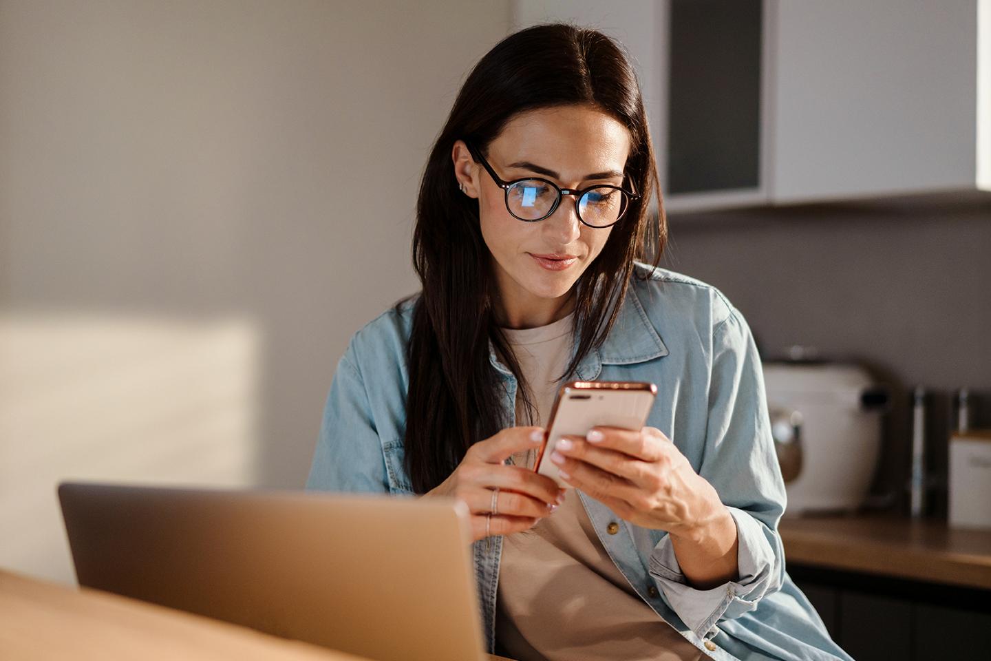 Une personne assise devant un ordinateur portable regarde son téléphone.