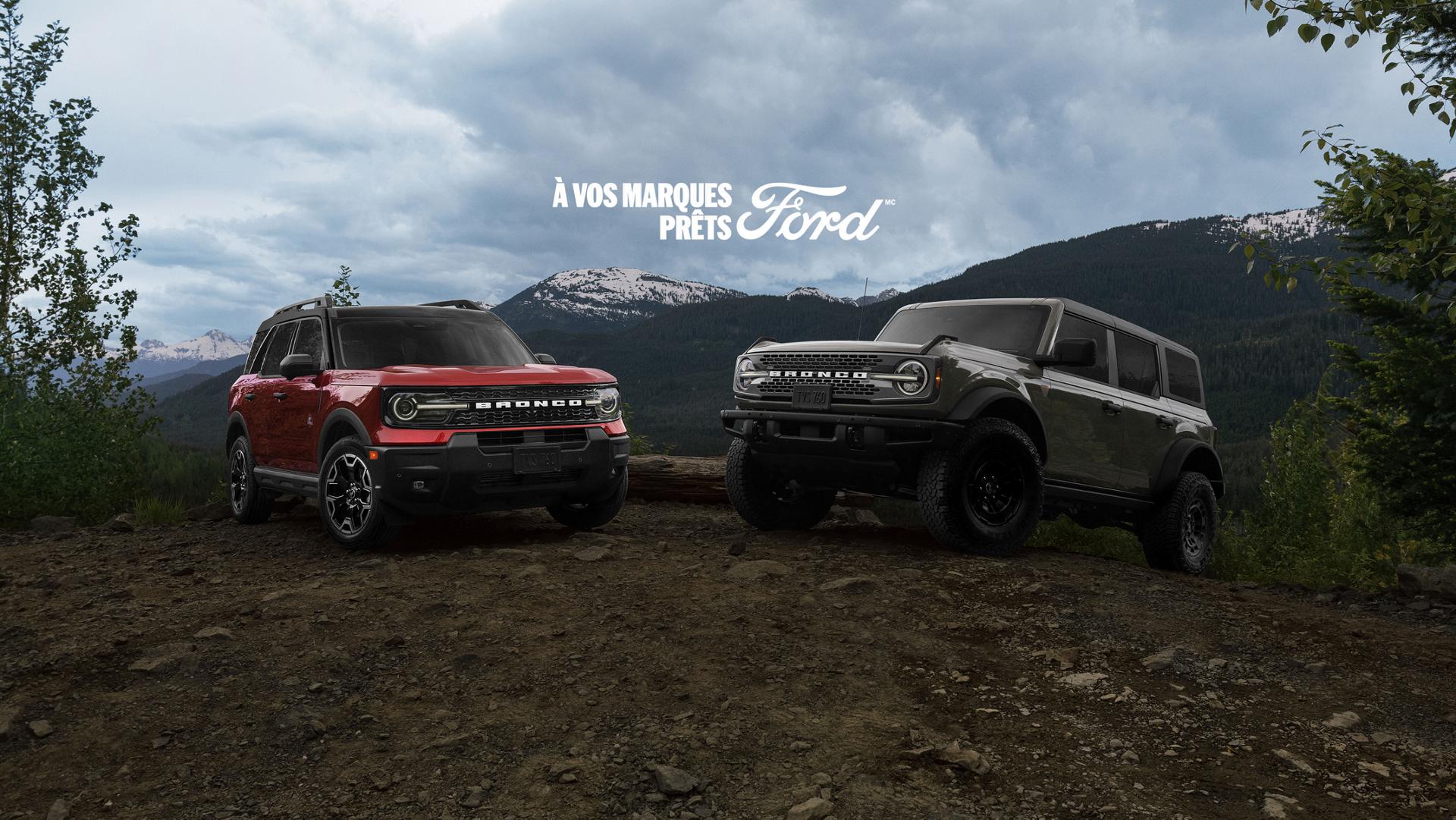 Un Ford Bronco Sport rouge à gauche et un Bronco gris à quatre portes à droite, garés sur une colline de terre battue avec les montagnes en arrière-plan et l'inscription « À vos marques prêts Ford » au-dessus d'eux.