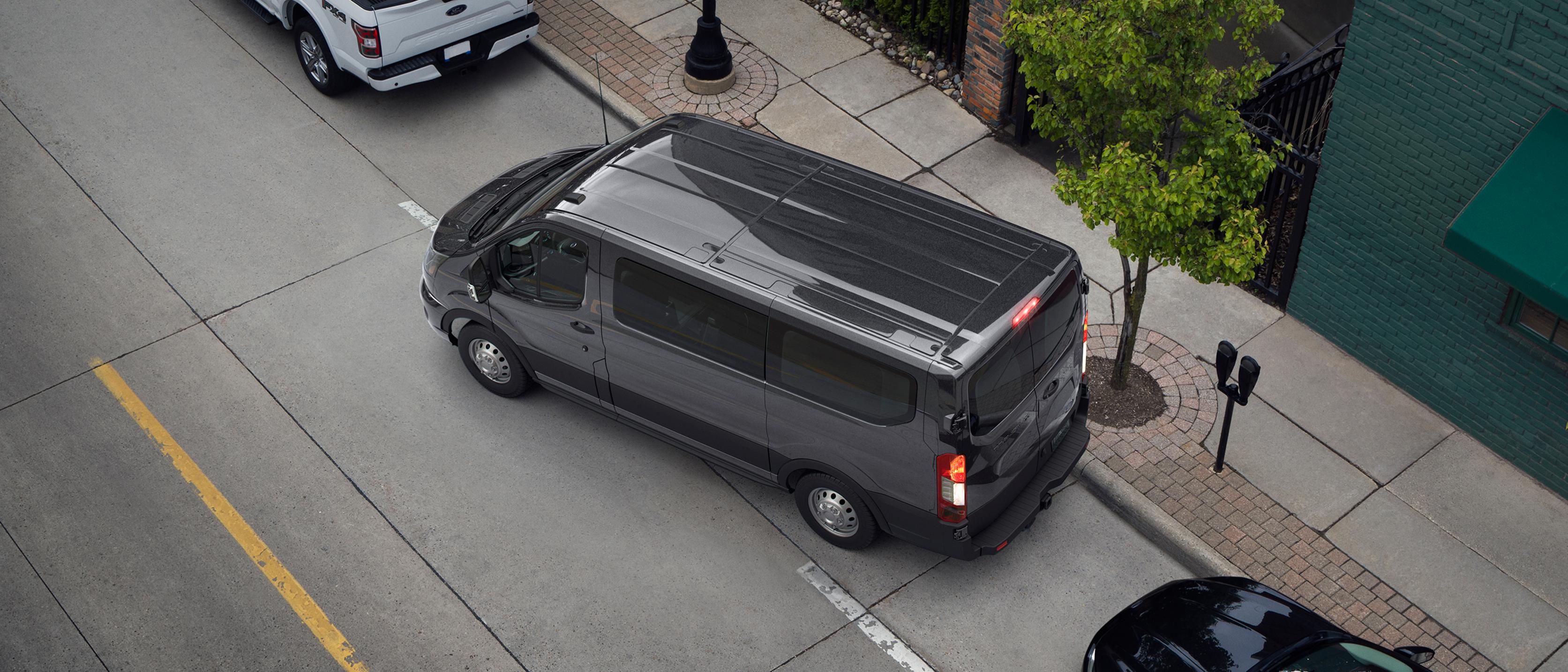 Vue au-dessus de la tête d’une fourgonnette Ford Transit® 2027 gris foncé effectuant un stationnement en parallèle dans une rue de la ville, entre d’autres véhicules