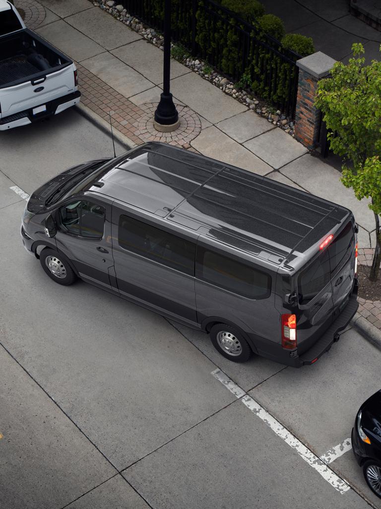 Vue aérienne d’une fourgonnette de transport de passagers Ford Transit® 2027 gris foncé effectuant un stationnement en parallèle dans une rue de la ville