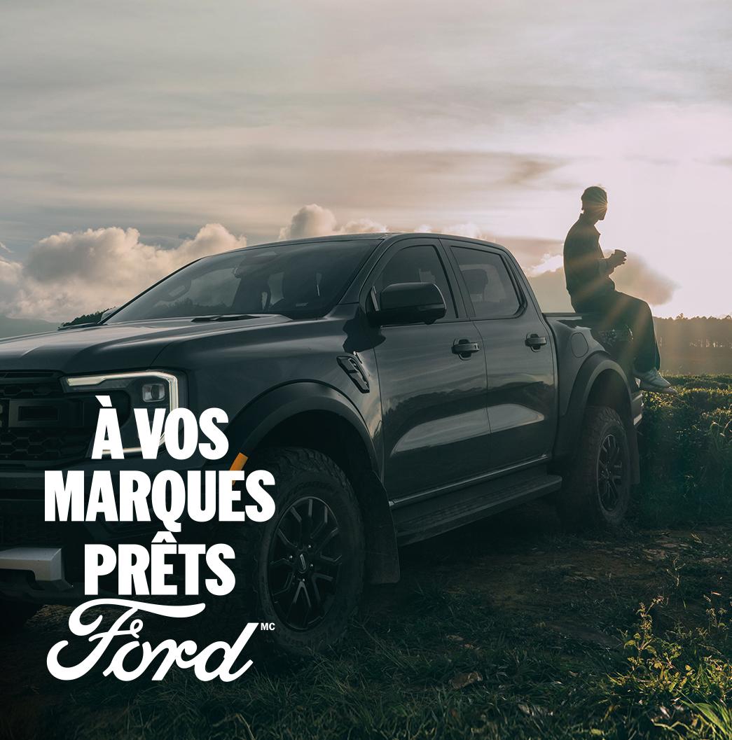 Vue de trois quarts d'un Ford Ranger Raptor gris garé sur l'herbe. Une personne est assise à l'arrière du pick-up, le soleil brille en arrière-plan. À vos marques, prêts, Ford.