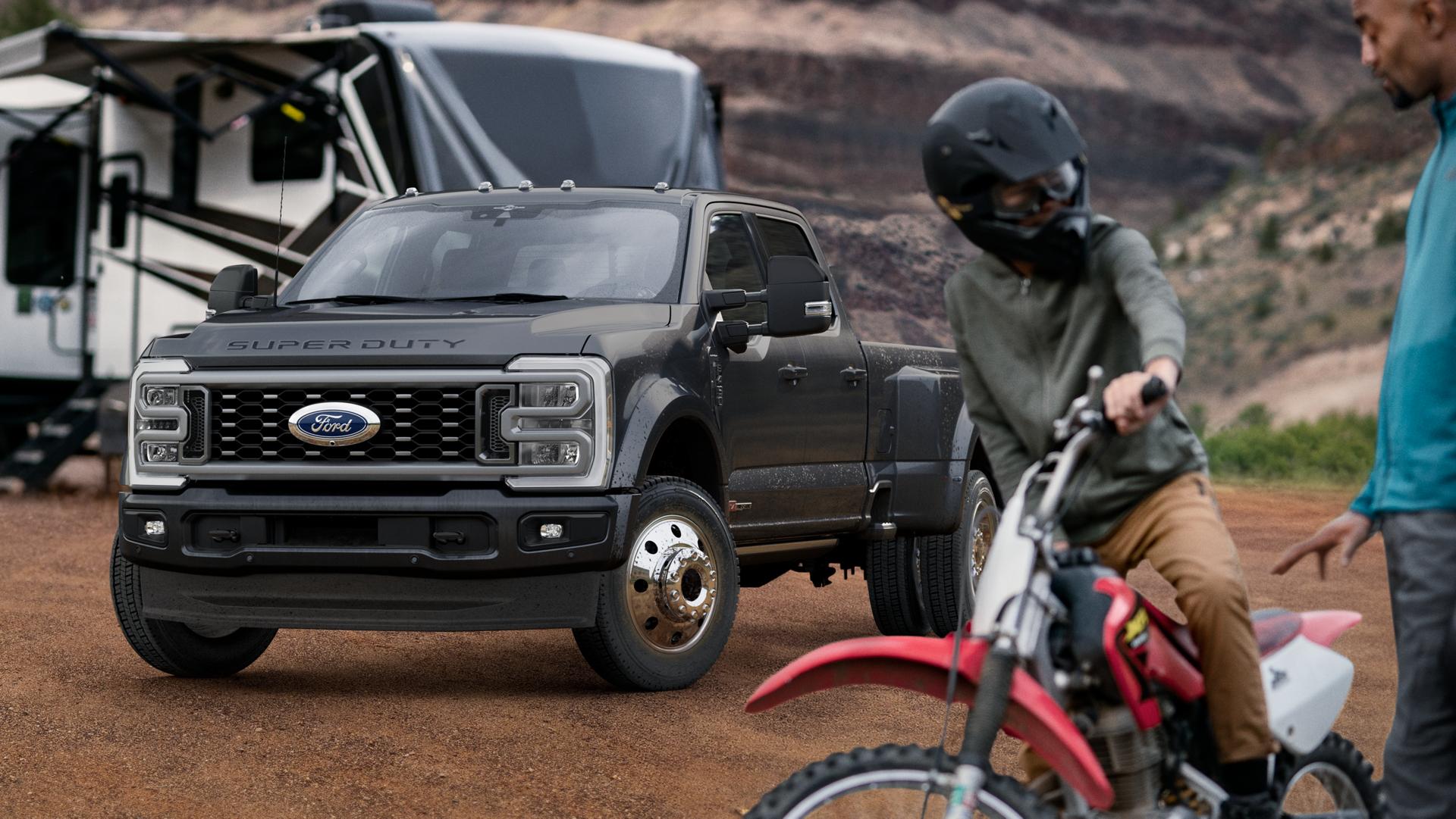 Modèle Ford Super Duty® F-450® Platinum® 2026 en gris carbonisé stationné sur un terrain de camping avec des caravanes en arrière-plan et une personne sur un vélo en avant-plan.
