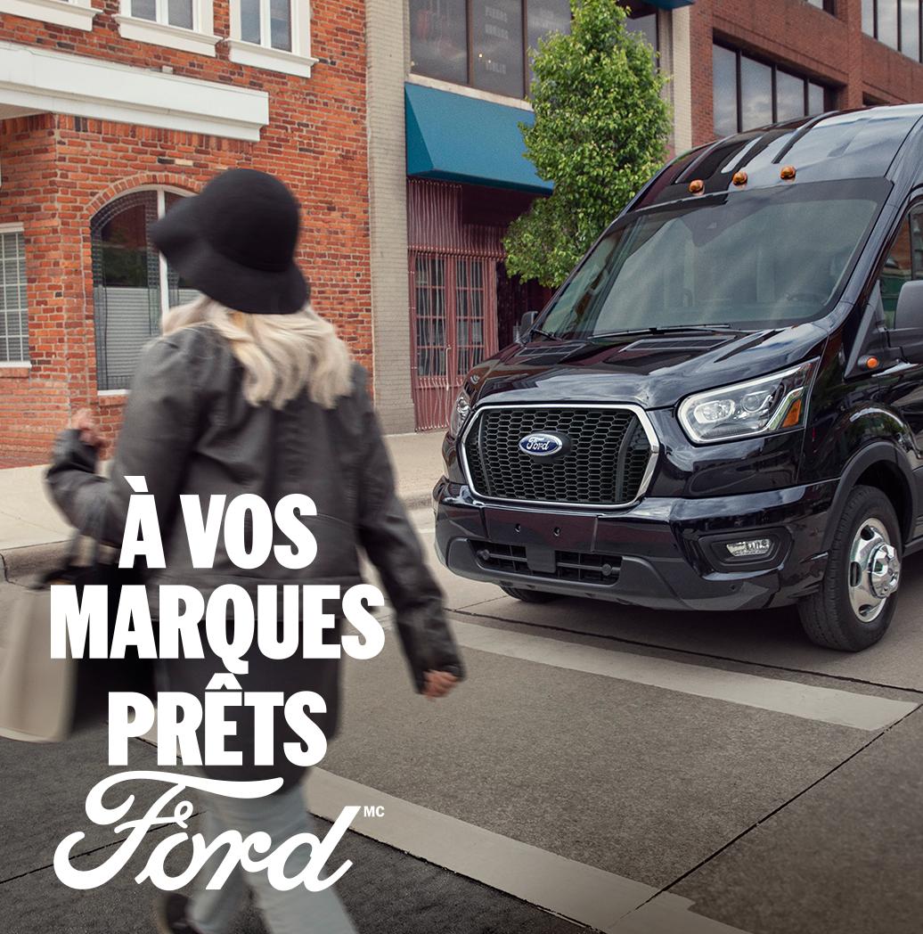 Vue de trois quarts avant d'un Ford Transit 2025 roulant en milieu urbain. À vos marques, prêts, Ford.