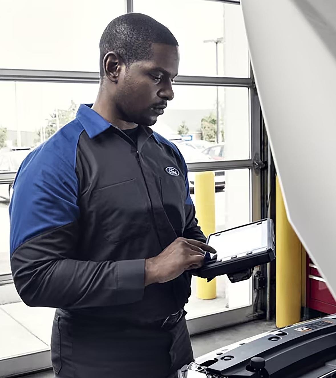 Un technicien à l’entretien de Ford fait une saisie sur une tablette tout en regardant sous le capot d’un véhicule Ford dans l’aire de service.