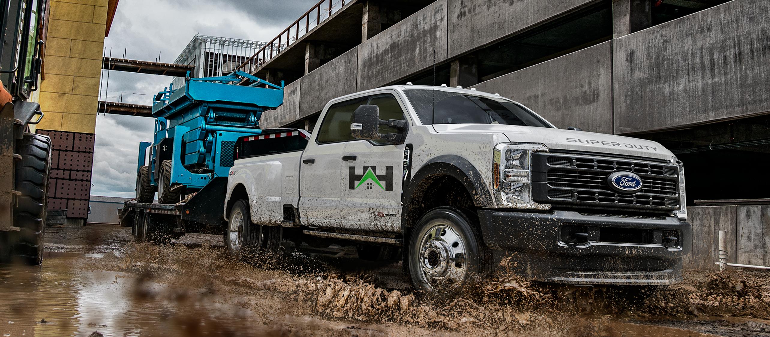 Modèle Ford Super Duty® F-450® XL 2026 tirant une remorque à plateforme avec un grand tracteur bleu dessus dans la boue et l’eau