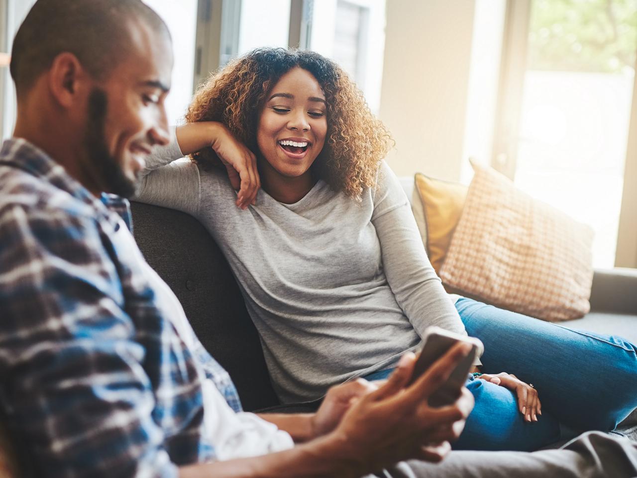 Un couple assis sur un canapé à la maison, souriant et regardant ensemble un téléphone intellig