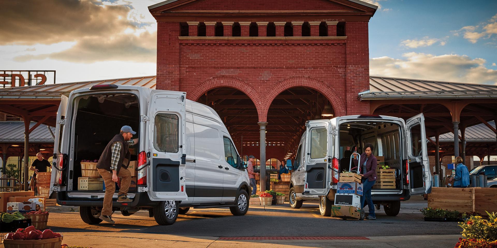 Two white Ford Transit® Vans with rear open and people unloading produce at a market