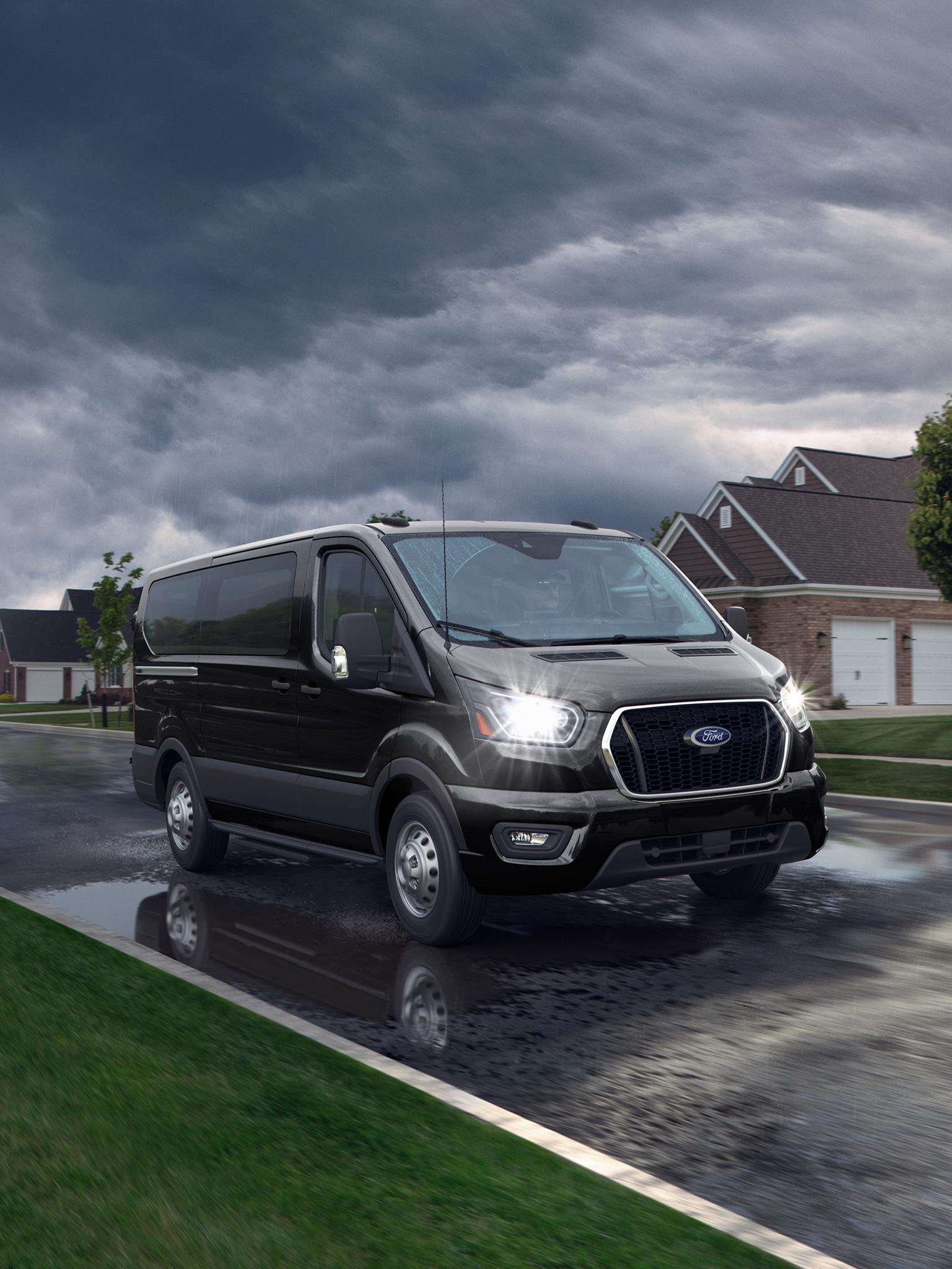 Une fourgonnette de transport de passagers Ford Transit® 2027 noire roule sur une rue résidentielle mouillée sous un ciel sombre et nuageux