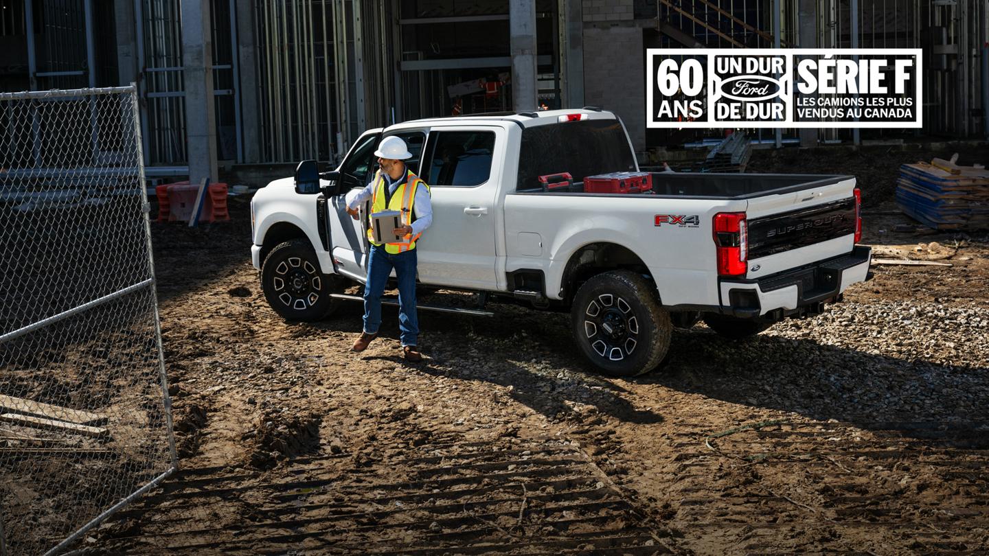 Ouvrier du bâtiment debout à côté d'un camion Ford Super Duty blanc vu de trois quarts arrière sur un chantier de construction. 60 ans – Ford un dur de dur – série F les camions les plus vendus au Canada 