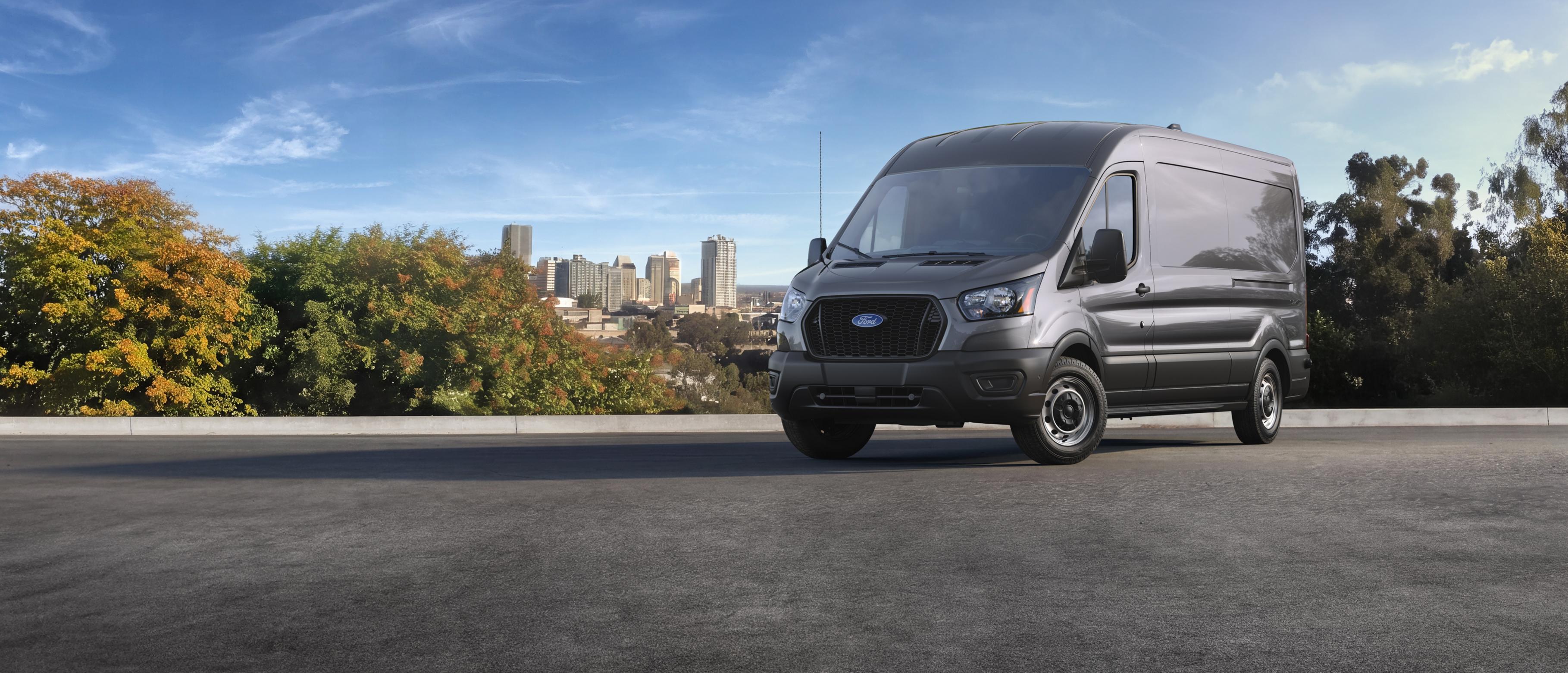 Fourgonnette utilitaire Ford Transit® 2027 grise roulant sur une route, avec une ligne d’horizon urbaine et des arbres en arrière-plan