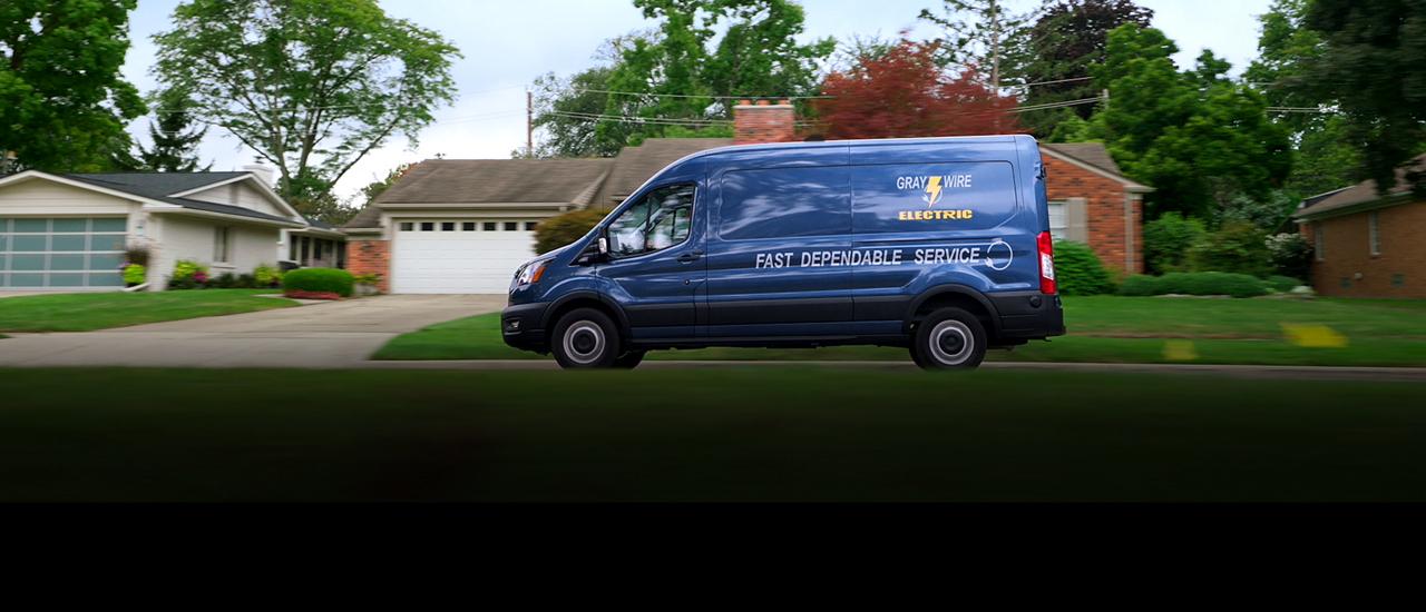 Une fourgonnette Ford Transit® 2027 bleue roulant devant des maisons dans un quartier résidentiel de banlieue