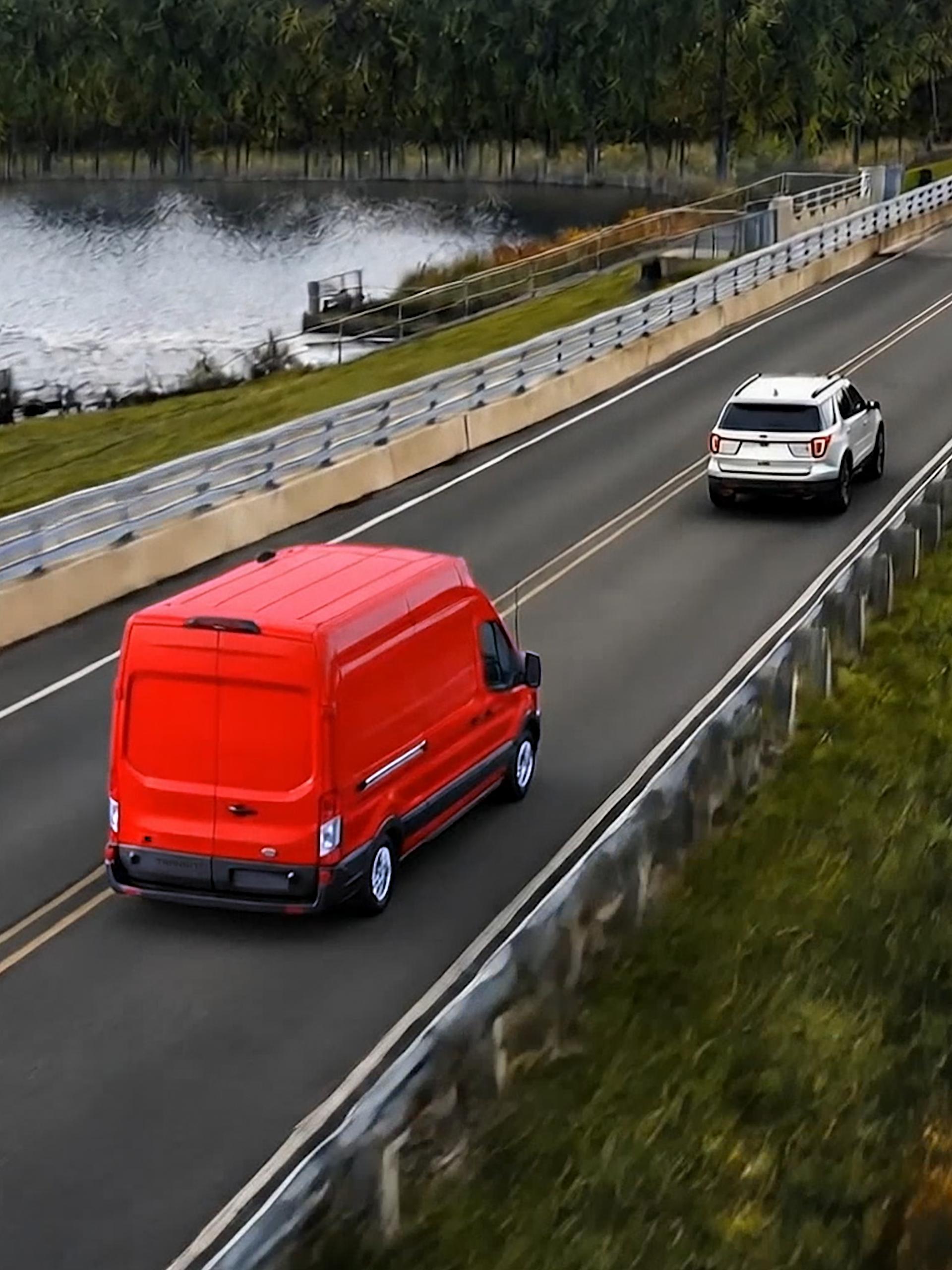 Fourgonnette Ford Transit® 2027 rouge roulant sur une route le long d’une rivière, avec un VUS blanc devant