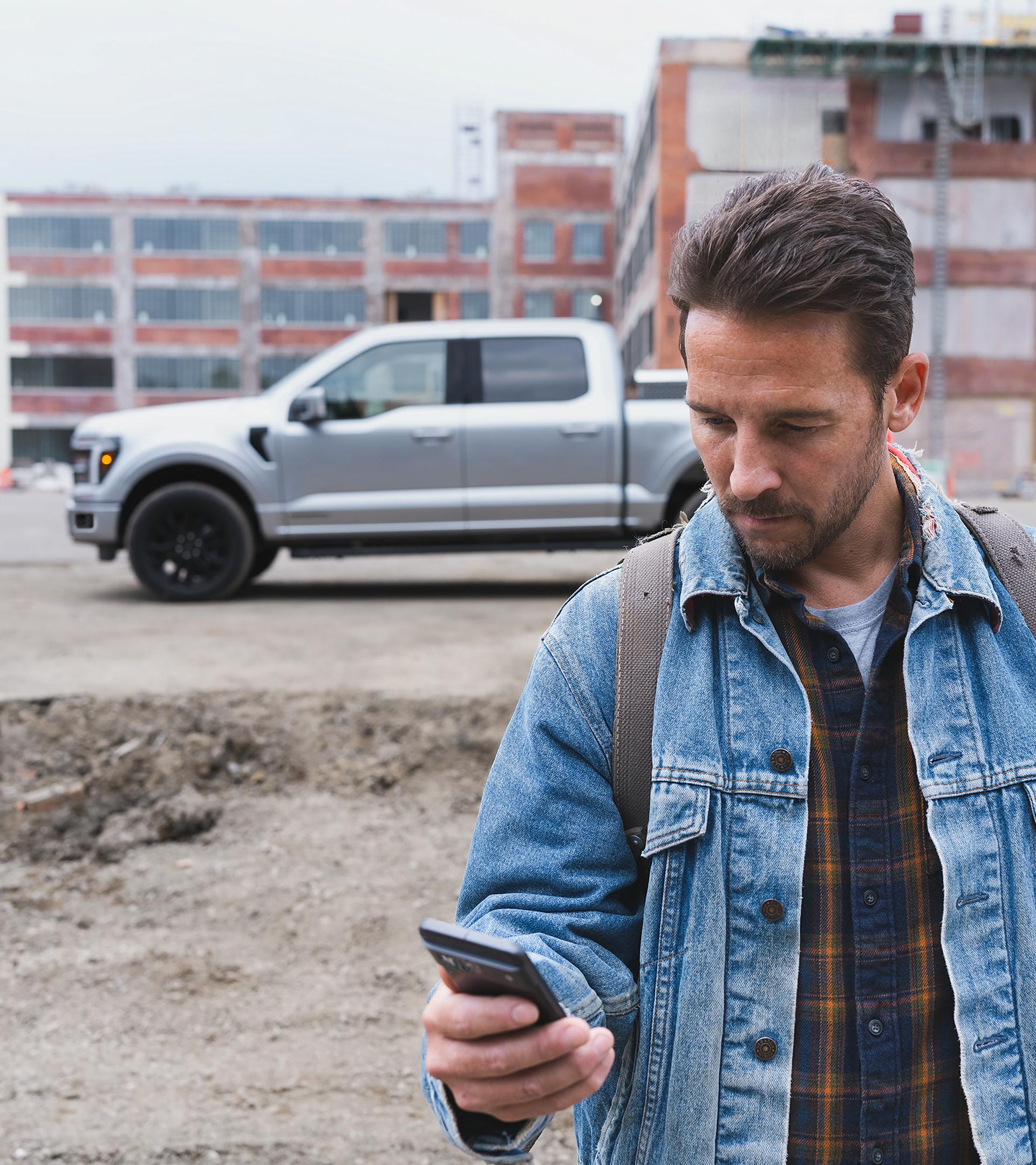 Un homme regardant son téléphone intelligent avec une camionnette F-150® en arrière-plan stationnée sur un chantier de construction
