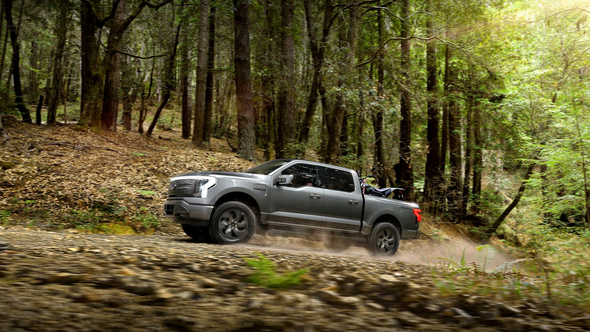 Un camion Ford F-150® Lightning® 2025 circulant sur une route poussiéreuse dans la forêt avec un VTT fixé dans la caisse de chargement