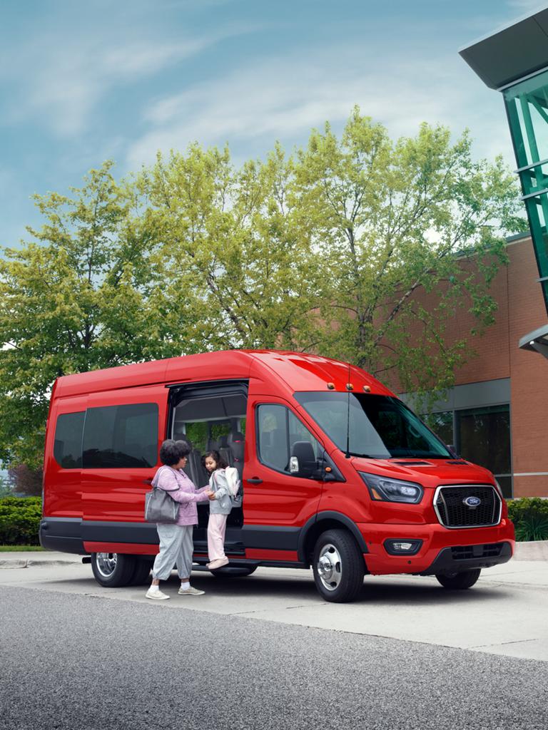 Une fourgonnette de transport de passagers Ford Transit® rouge stationnée devant un immeuble moderne, avec une famille à proximité