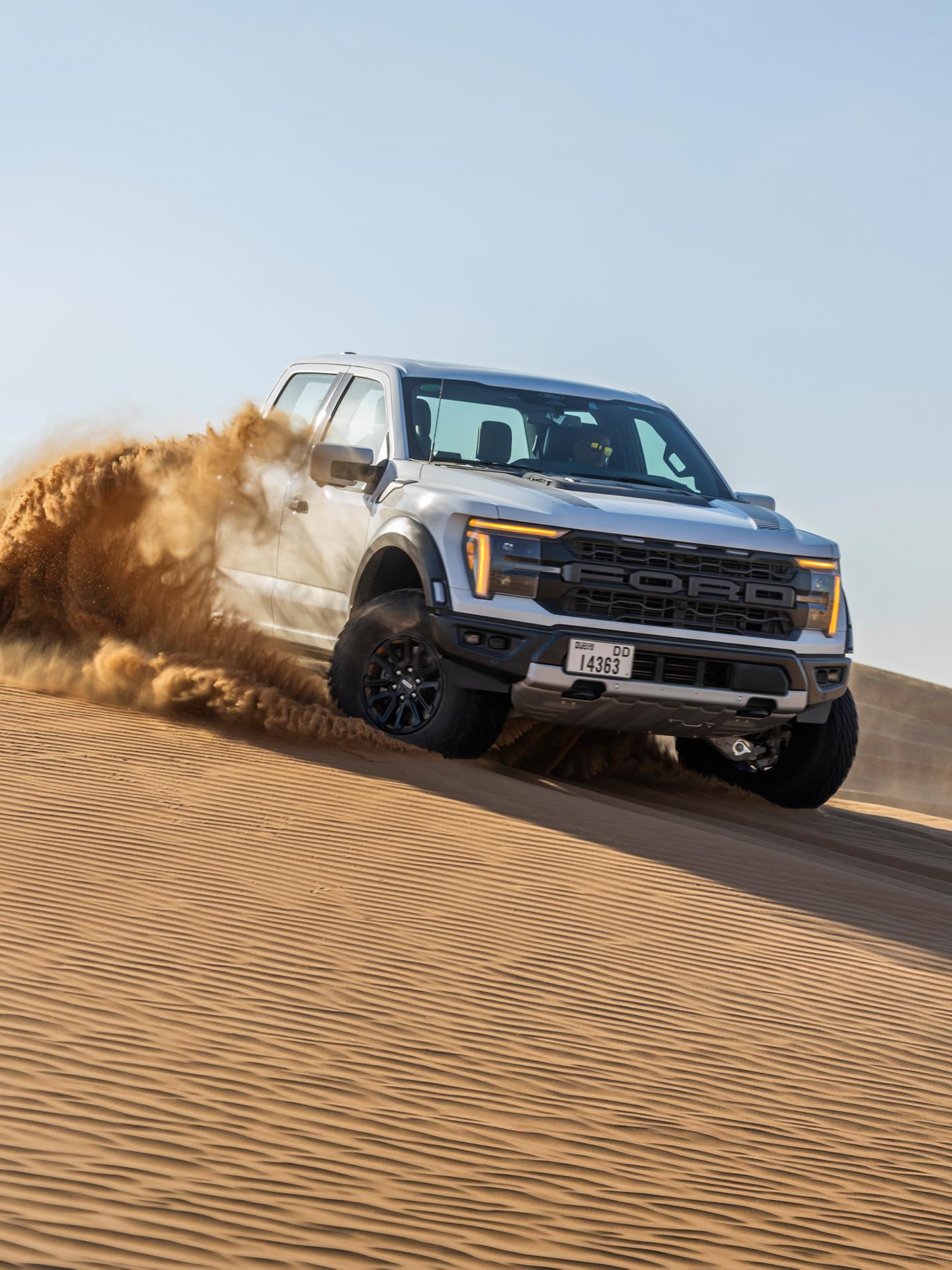 Un Ford F-150 Raptor bleu foncé roulant dans le sable et la poussière sur une dune du désert.