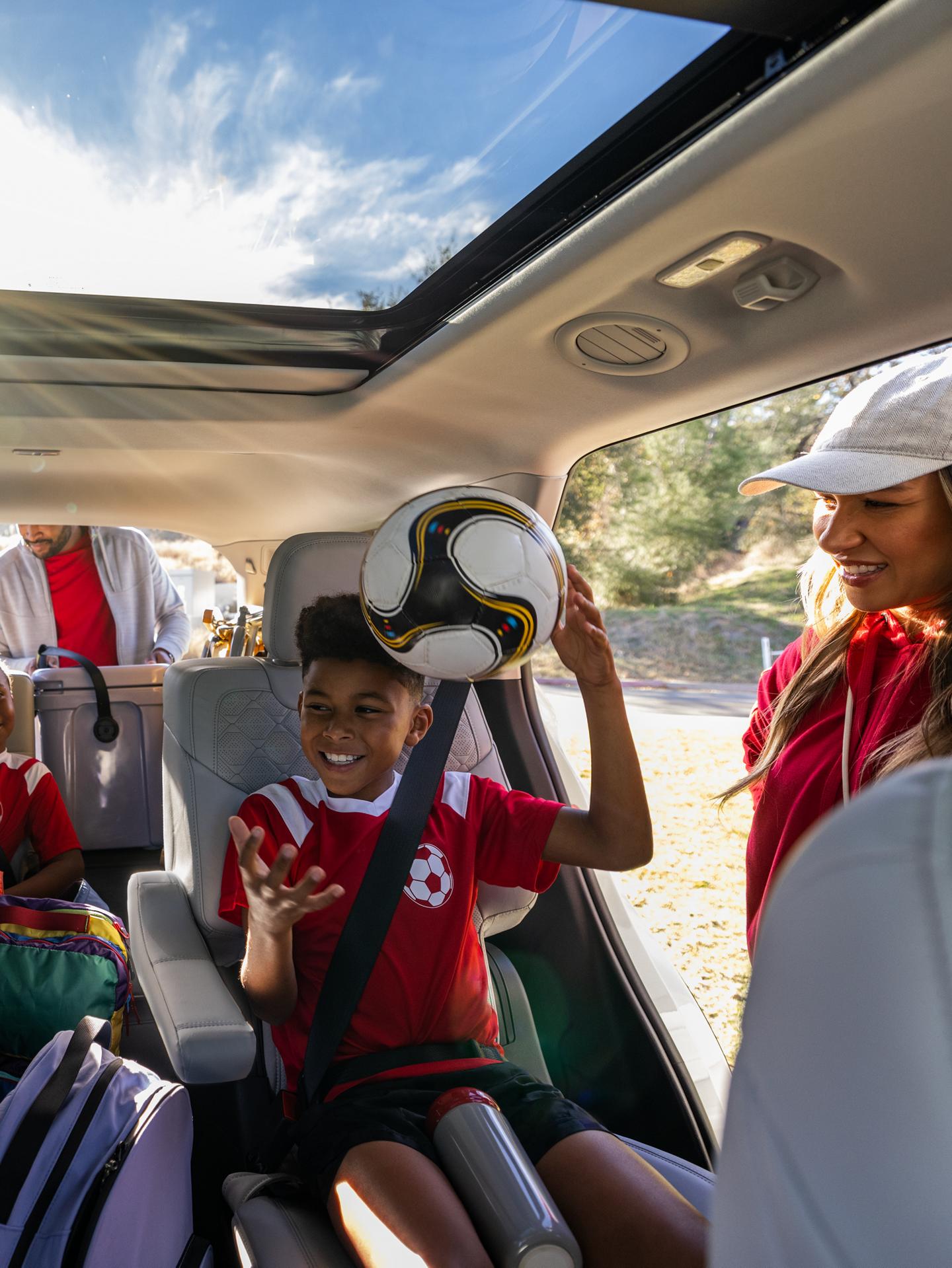 Un enfant attaché sur la banquette arrière d’un Ford tient un ballon de soccer, tandis que deux adultes s’affairent autour du véhicule.