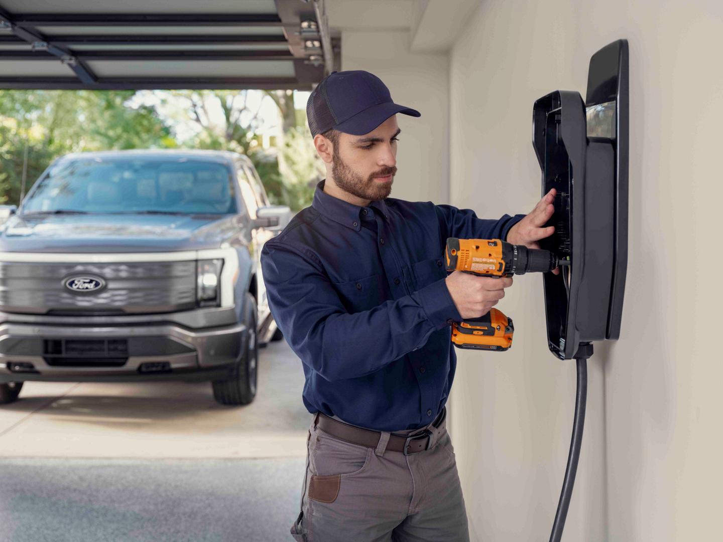 Un homme installant un poste de recharge à domicile dans un garage avec un camion Ford F-150® Lightning® 2025 stationné à l’extérieur de la porte