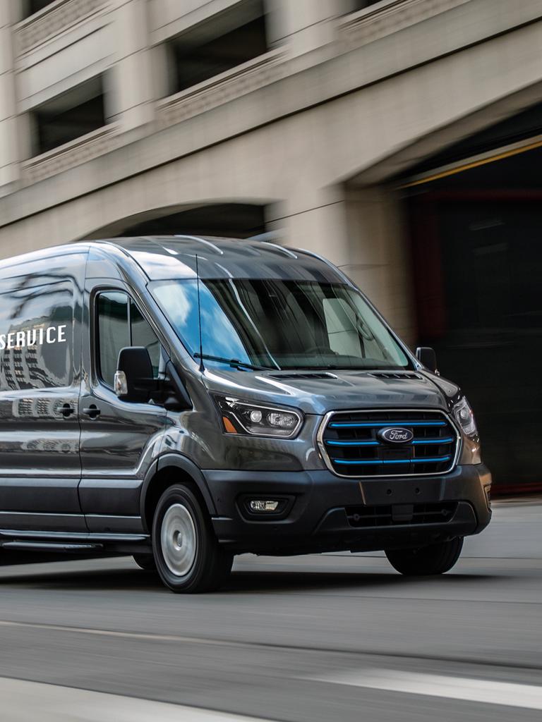 Fourgonnette utilitaire Ford E-Transit™ 2027 gris foncé avec des accents bleus sur la calandre, roulant dans une rue de la ville le long d’un immeuble moderne