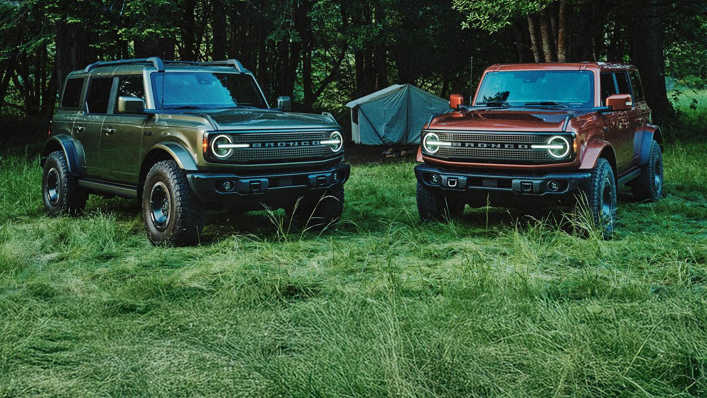 Deux VUS Ford Bronco garés côte à côte, en vue de trois quarts avant, dans un terrain de camping boisé. Un Bronco vert foncé se trouve à gauche, et un Bronco rouge cuivré à droite.