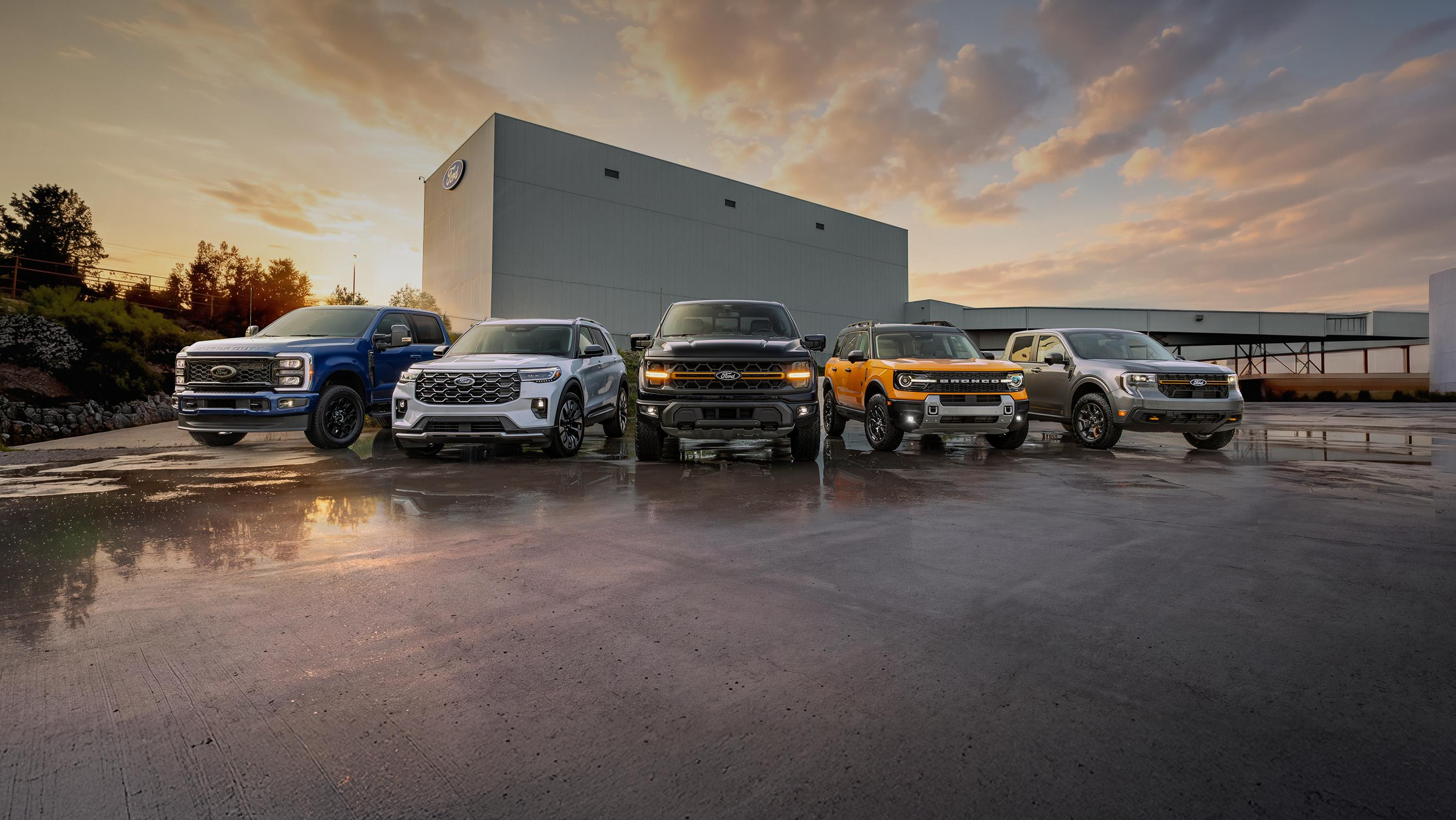 Une gamme de véhicules Ford est stationnée devant une usine Ford, sur un asphalte détrempé par la pluie, sous un ciel nuageux au coucher du soleil. Le texte indique : Obtenez de belles offres sur les Ford neufs