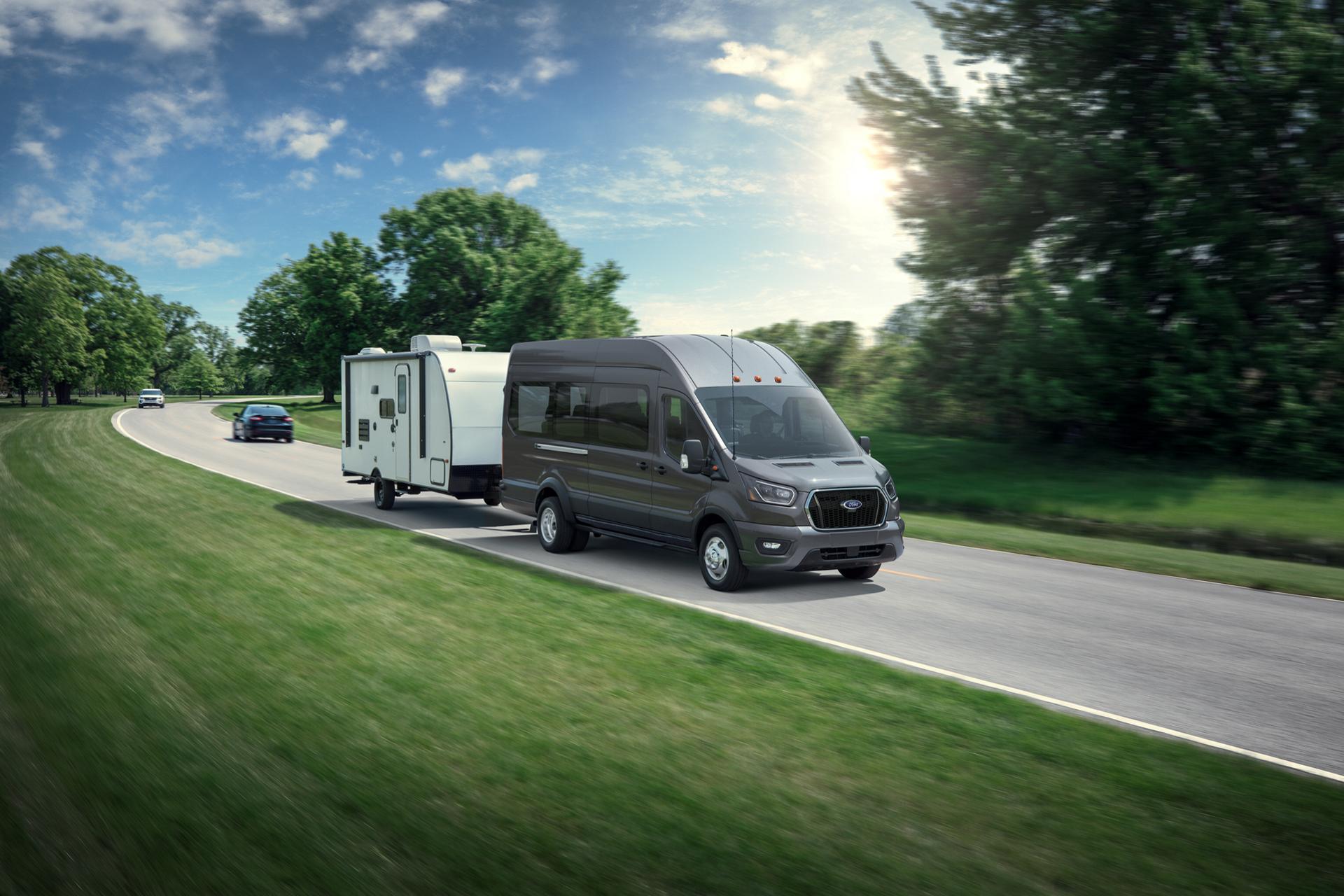 Une fourgonnette Ford Transit® 2027 blanche tirant une petite remorque sur une route sinueuse avec des arbres