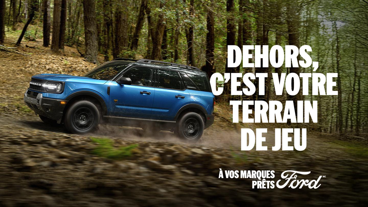 Vue de profil d'un Bronco Sport bleu roulant hors route sur un chemin de terre à travers une forêt. Dehors, c'est votre terrain de jeu. À vos marques, prêts, Ford.