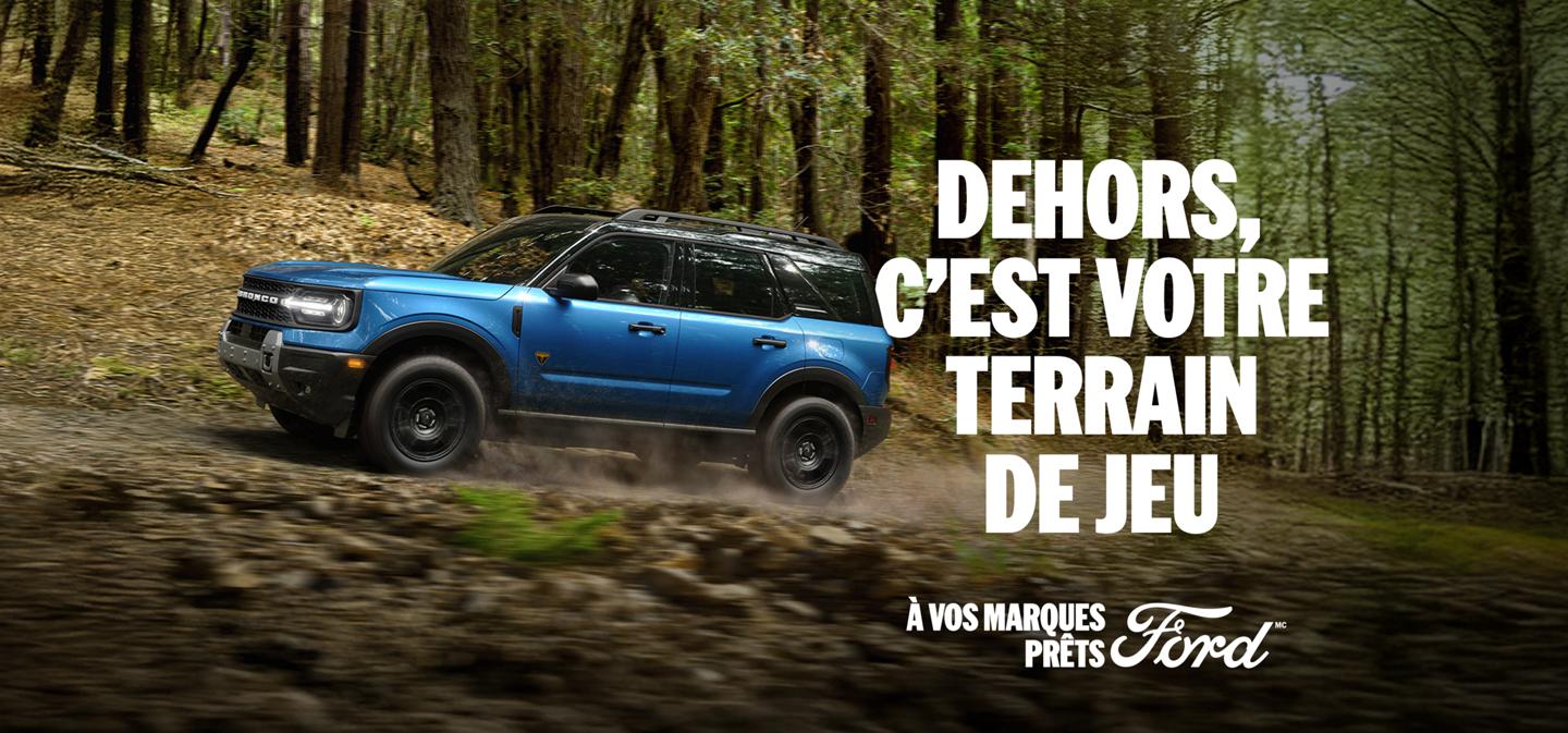 Vue de profil d'un Bronco Sport bleu roulant hors route sur un chemin de terre à travers une forêt. Dehors, c'est votre terrain de jeu. À vos marques, prêts, Ford.