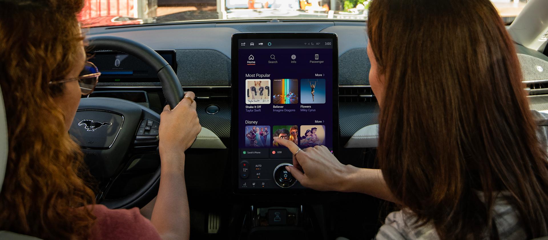 Deux femmes assises sur le siège avant d’un VUS Mustang Mach-E® 2026 avec le passager naviguant parmi les options de karaoké dans le forfait de connectivité Ford en option