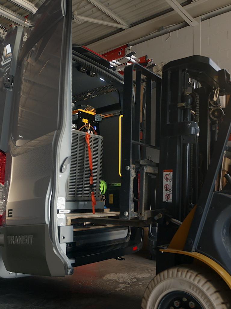 Un chariot élévateur charge des sacs à l’arrière d’une fourgonnette utilitaire Ford E-Transit™ 2027 rouge aux portes ouvertes, stationnée devant un bâtiment