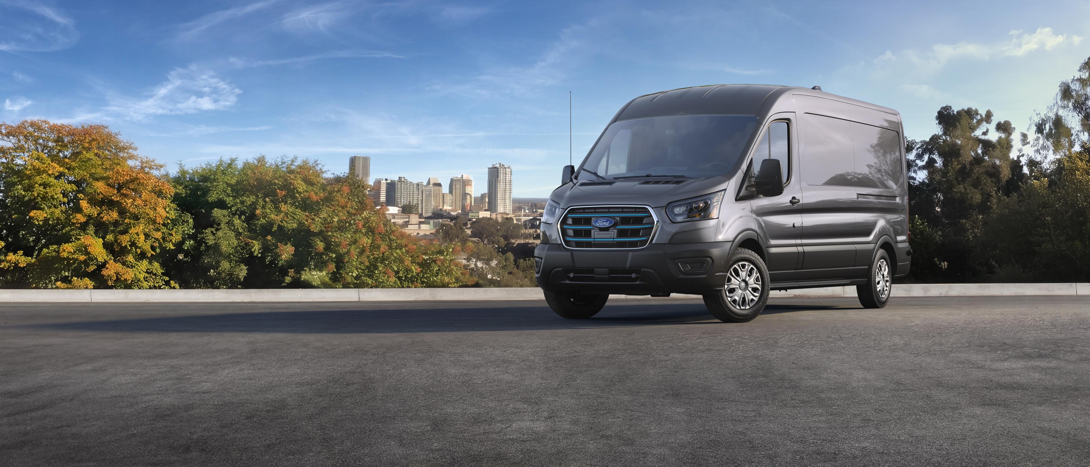 Une fourgonnette utilitaire Ford E-Transit™ 2027 grise stationnée sur une route asphaltée, avec une ligne d’horizon urbaine et des arbres en arrière-plan sous un ciel dégagé