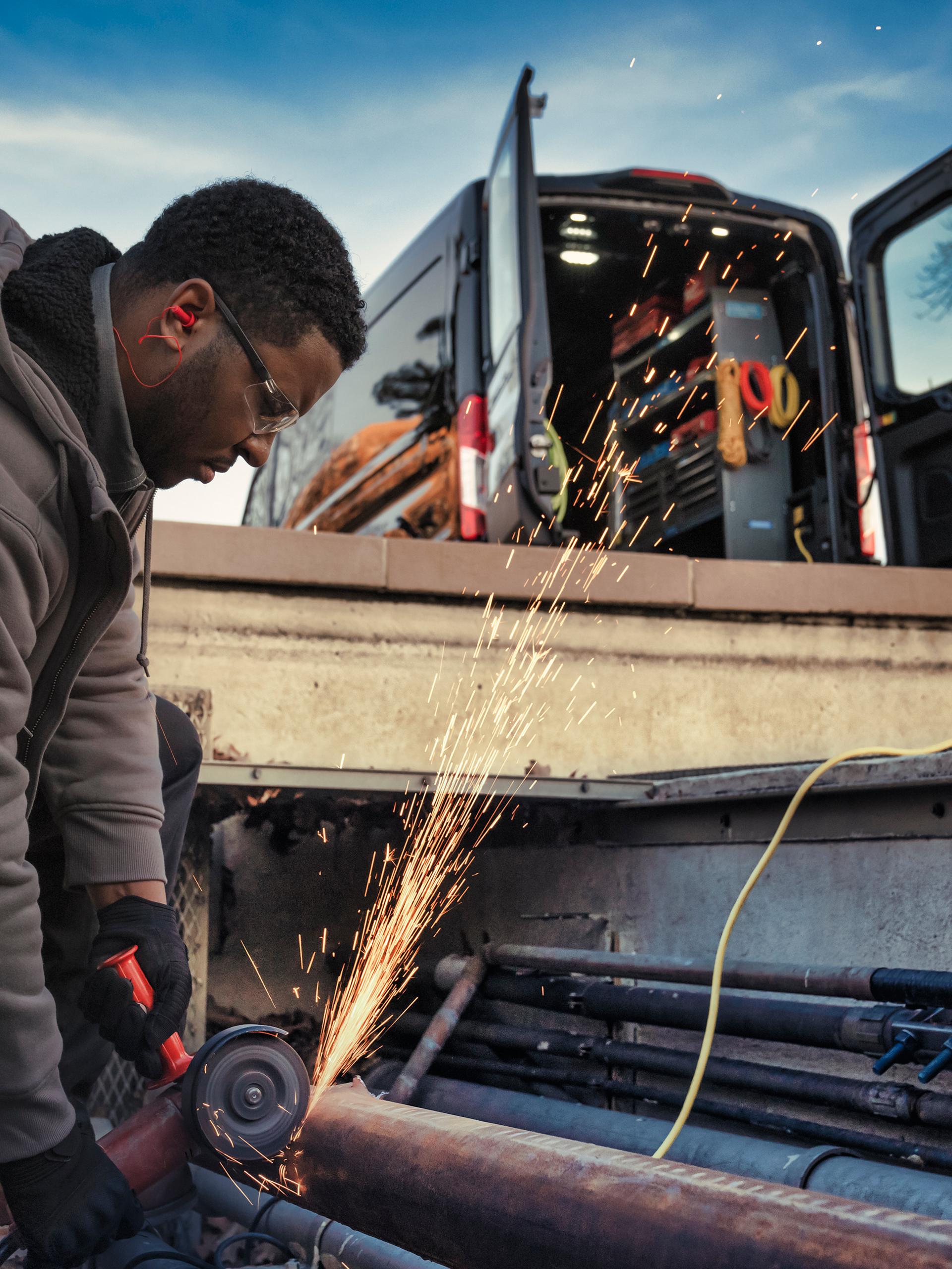 Travailleur utilisant une meuleuse, avec des étincelles qui jaillissent, et une fourgonnette Ford Transit® 2027 aux portes ouvertes en arrière-plan