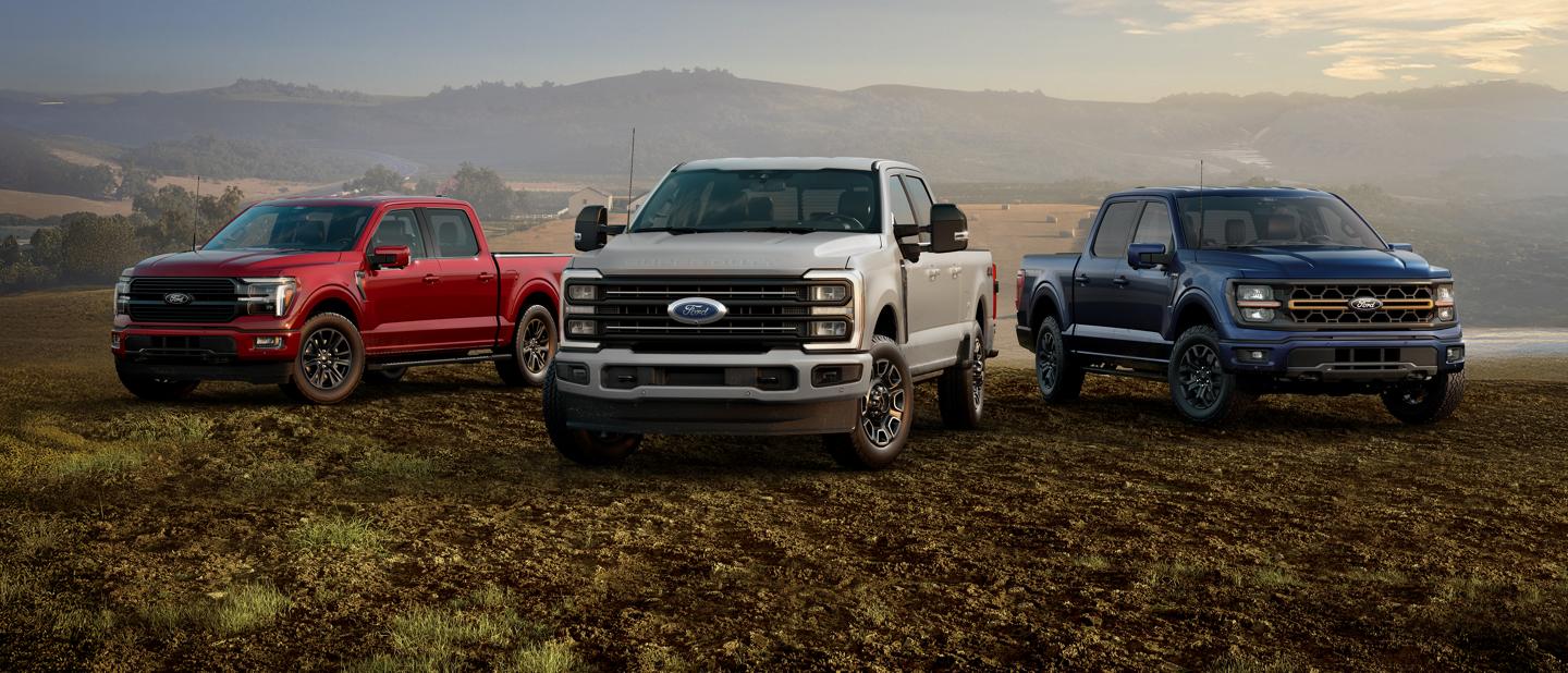 Trois camions Ford de la série F, rouge, blanc et bleu, garés côte à côte dans un champ, avec des collines ondulantes en arrière-plan.
