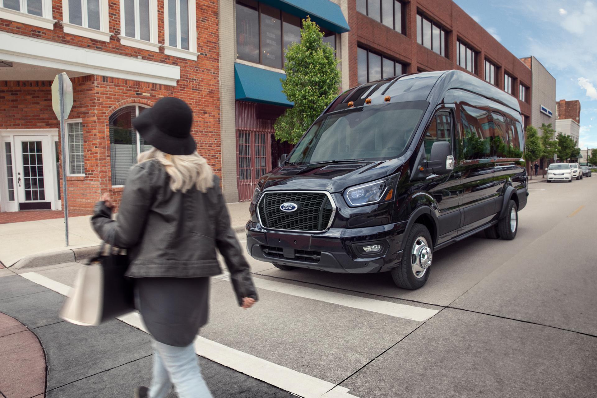 Une femme marchant devant une fourgonnette Ford Transit® 2027 noire stationnée dans une rue de la ville bordée d’immeubles en briques